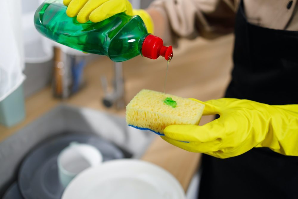 Close up of woman applying dishes soap liquid from cleanser bottle to sponge at kitchen